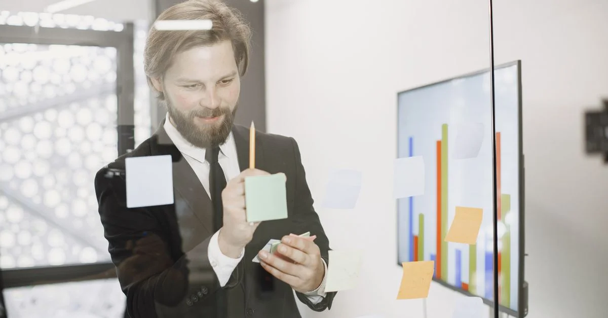 Homme professionnel en costume utilisant des post-it sur un mur en verre pour un brainstorming d'affaires.