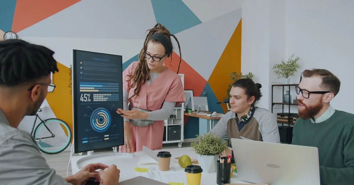 Dans un environnement de bureau dynamique, une équipe aux profils variés discute des analyses affichées sur un écran, favorisant ainsi la collaboration.