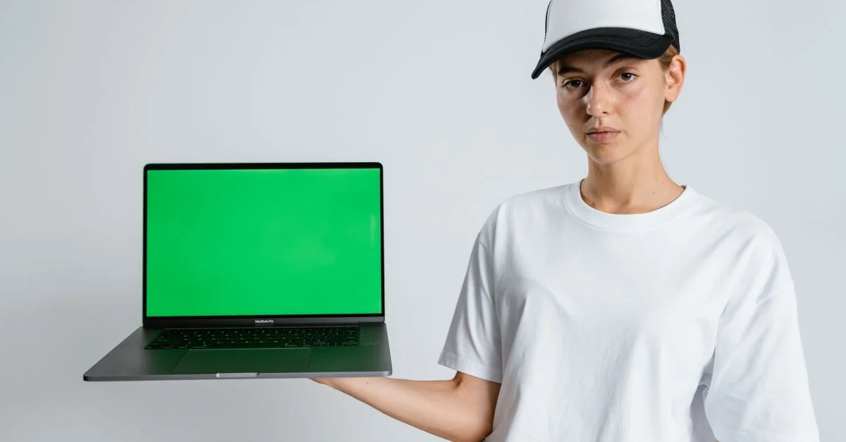 Portrait d'une personne en casquette tenant un ordinateur portable avec un écran vert sur un fond neutre.