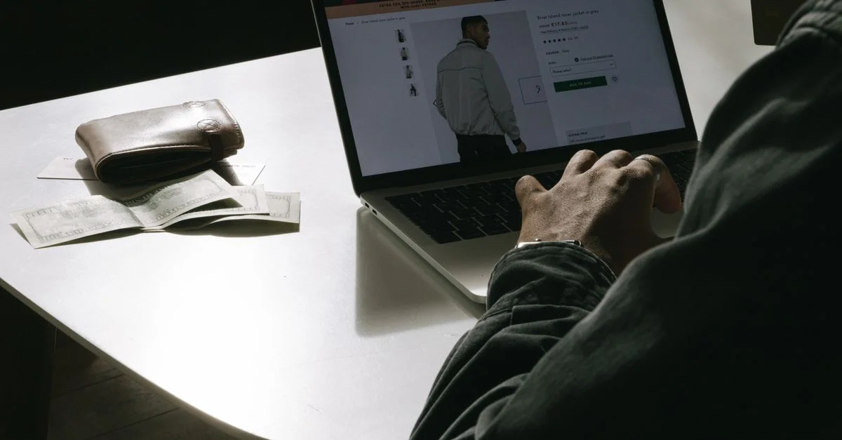 Un homme adulte achète des vêtements en ligne chez lui en utilisant un ordinateur portable, avec un portefeuille sur la table.
